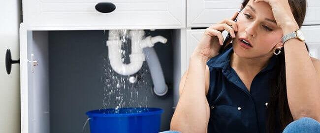 woman beside leaking pipe under sink calling emergency plumber